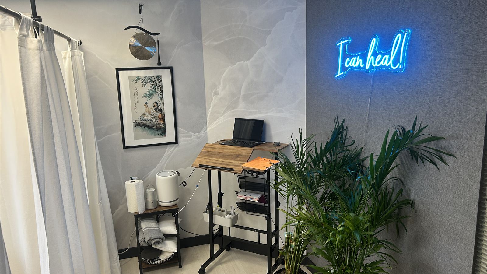 A treatment corner at Aculosophy with white curtains, a standing desk, a Chinese painting and an I can heal! neon sign